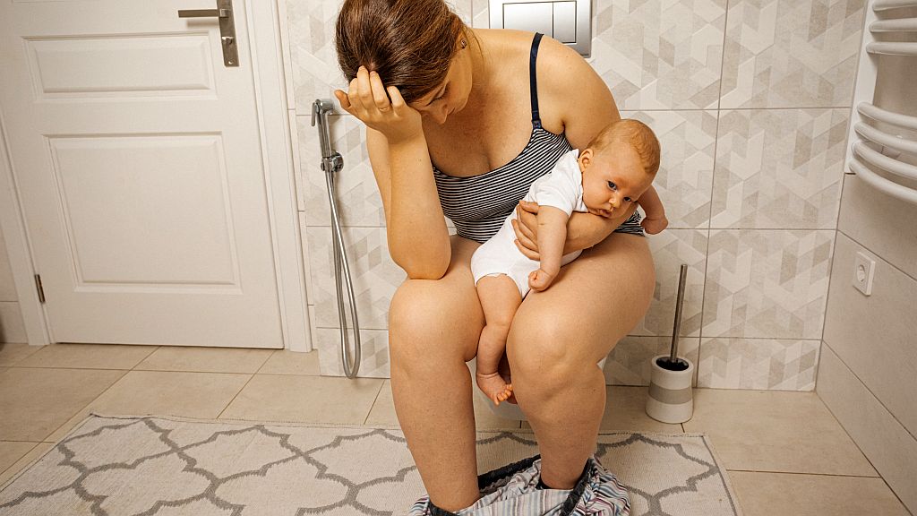 Frau mit Hämorrhoiden nach der Geburt, mit Baby im Arm, gestresster Ausdruck, auf der Toilette sitzend