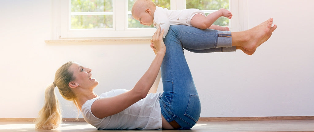 Mutter auf dem Rücken liegend hält ihr Baby, das auf ihren angewinkelten Beinen über ihrem Körper liegt, an den Händen, als Beckenbodentraining nach der Geburt.