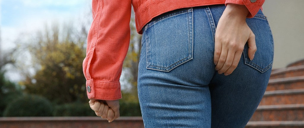 Symbolisches Bild für ein Hämorrhoiden Problem: Foto vom Unterkörper einer Frau von hinten im Alltag, in Jeans und roter kurzer Jacke, sie fasst sich an das Gesäß als Zeichen für Beschwerden durch Hämorrrhoiden