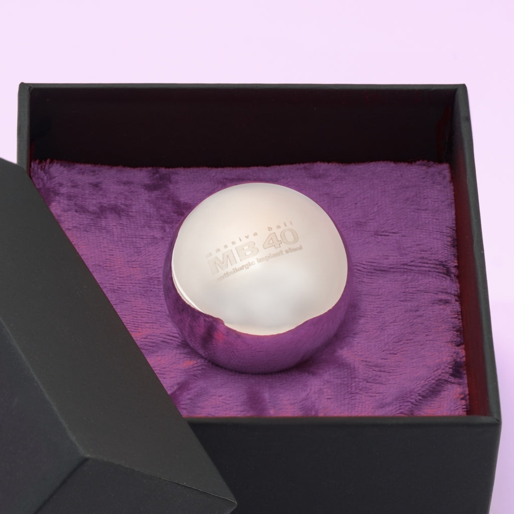 Theros® Massive Ball MB40 – esfera maciza de 40 mm de acero inoxidable antialérgico para mujeres y hombres para el entrenamiento del suelo pélvico en posiciones horizontales o para la estimulación de la próstata en una caja negra sobre terciopelo violeta oscuro, vista detallada.