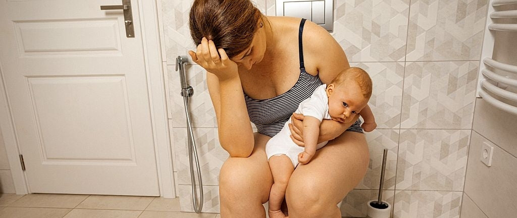 Mujer con hemorroides después del parto, con un bebé en brazos, expresión estresada, sentada en el inodoro